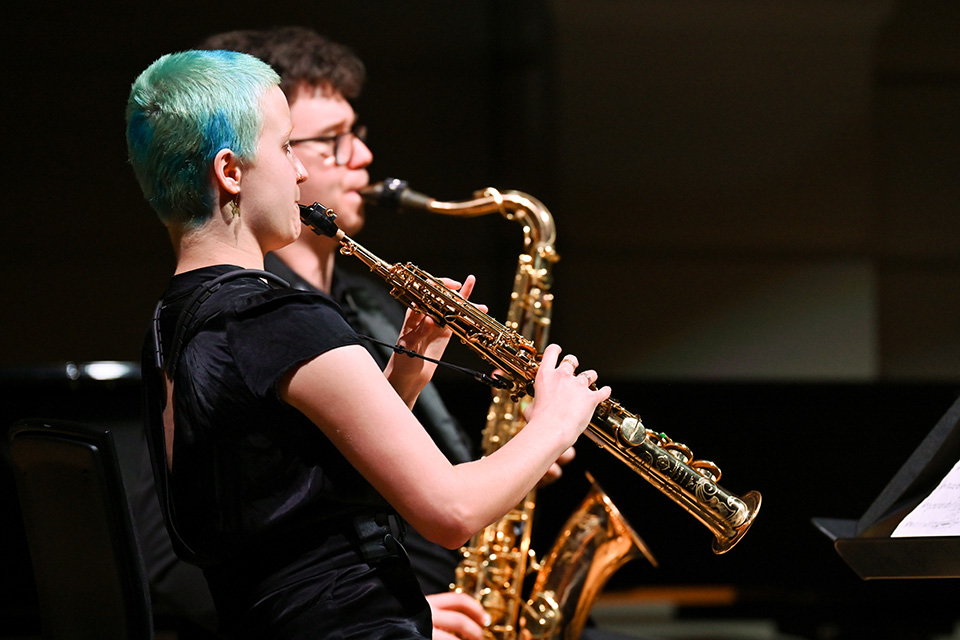 Two saxophonists performing in a chamber concert, on a dark stage.